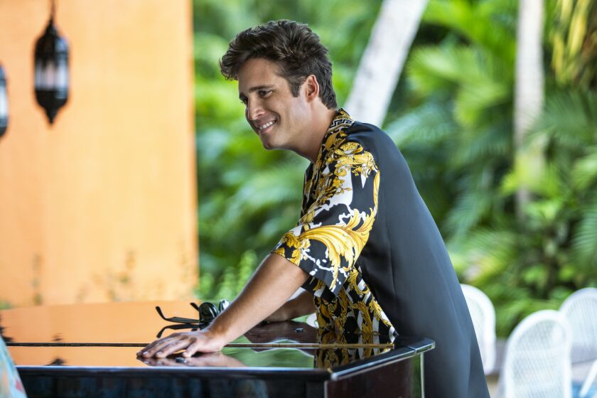 A man in a gold and black shirt leans on a black piano
