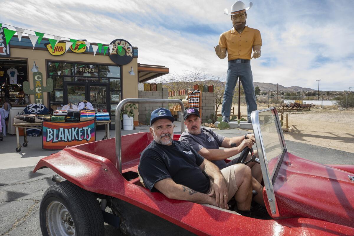 Two men sit in a red dune buggy outside a store, overlooked by a tall fiberglass figure of a man.