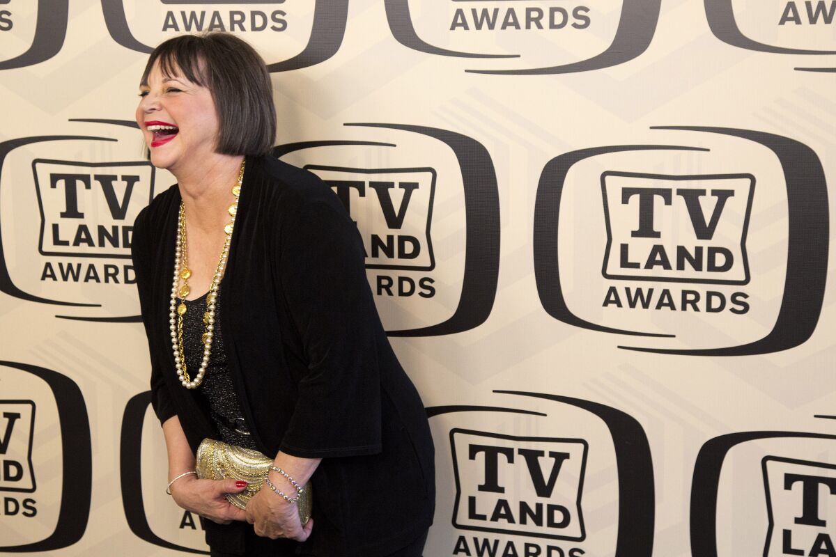 A woman with a dark bob haircut wearing long gold necklaces and a black outfit leans over and laughs