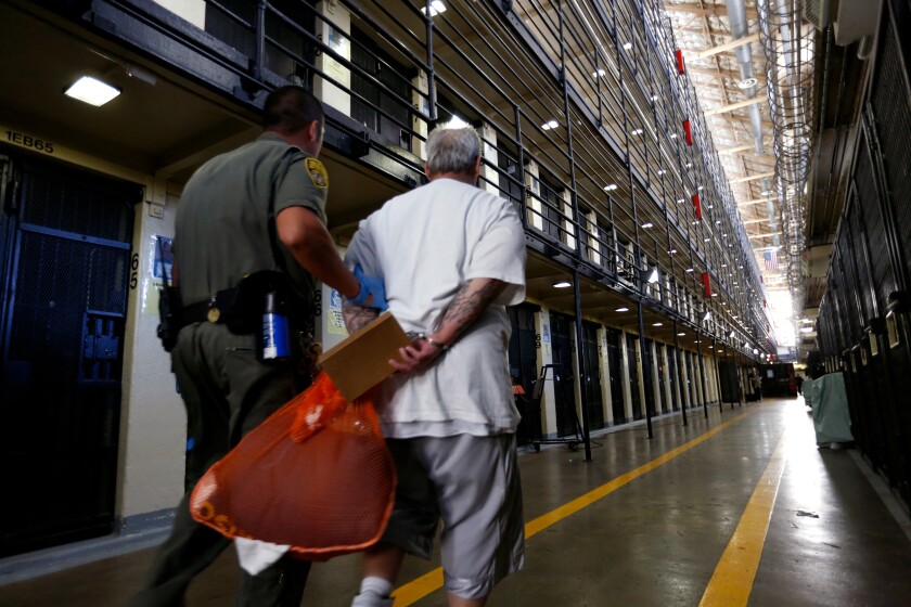 A guard escorts a prisoner carrying an orange bag between rows of cells