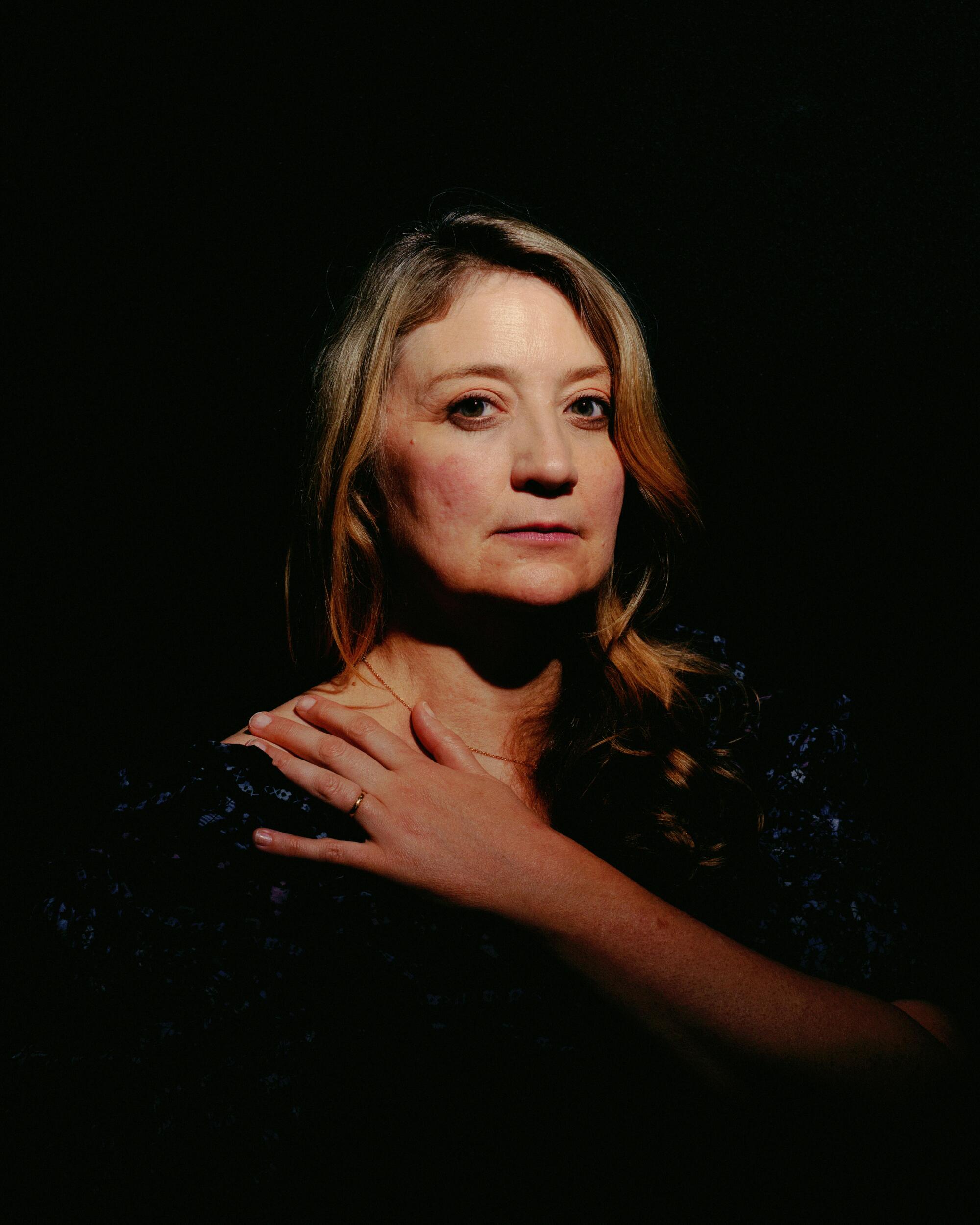 A woman rests her hand on her opposite shoulder surrounded by darkness