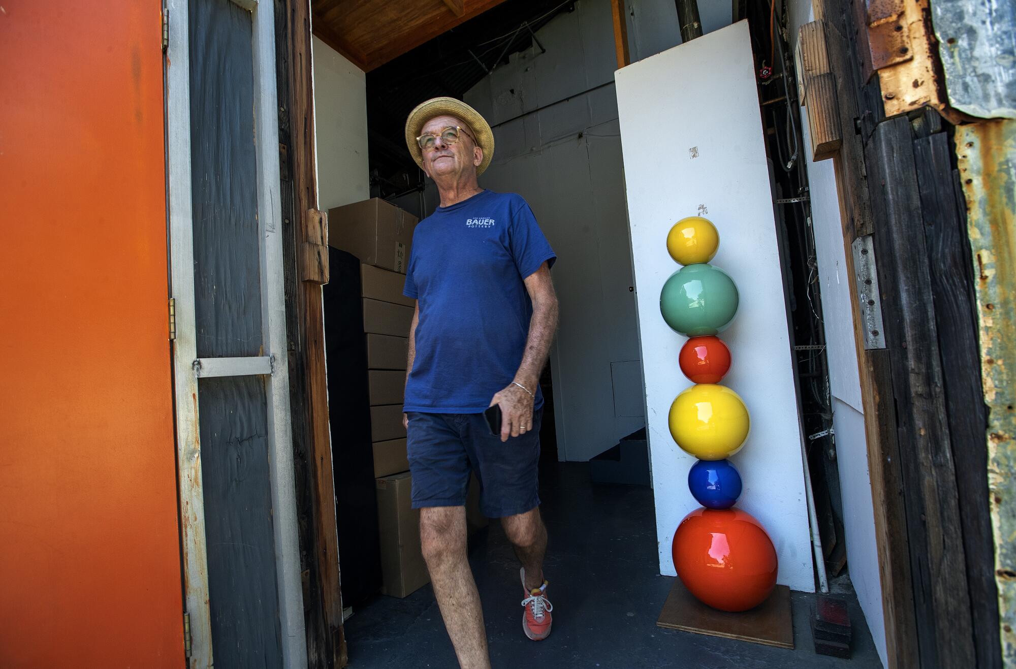 A man in a hat walks out a door next to a colorful tower of pottery balls