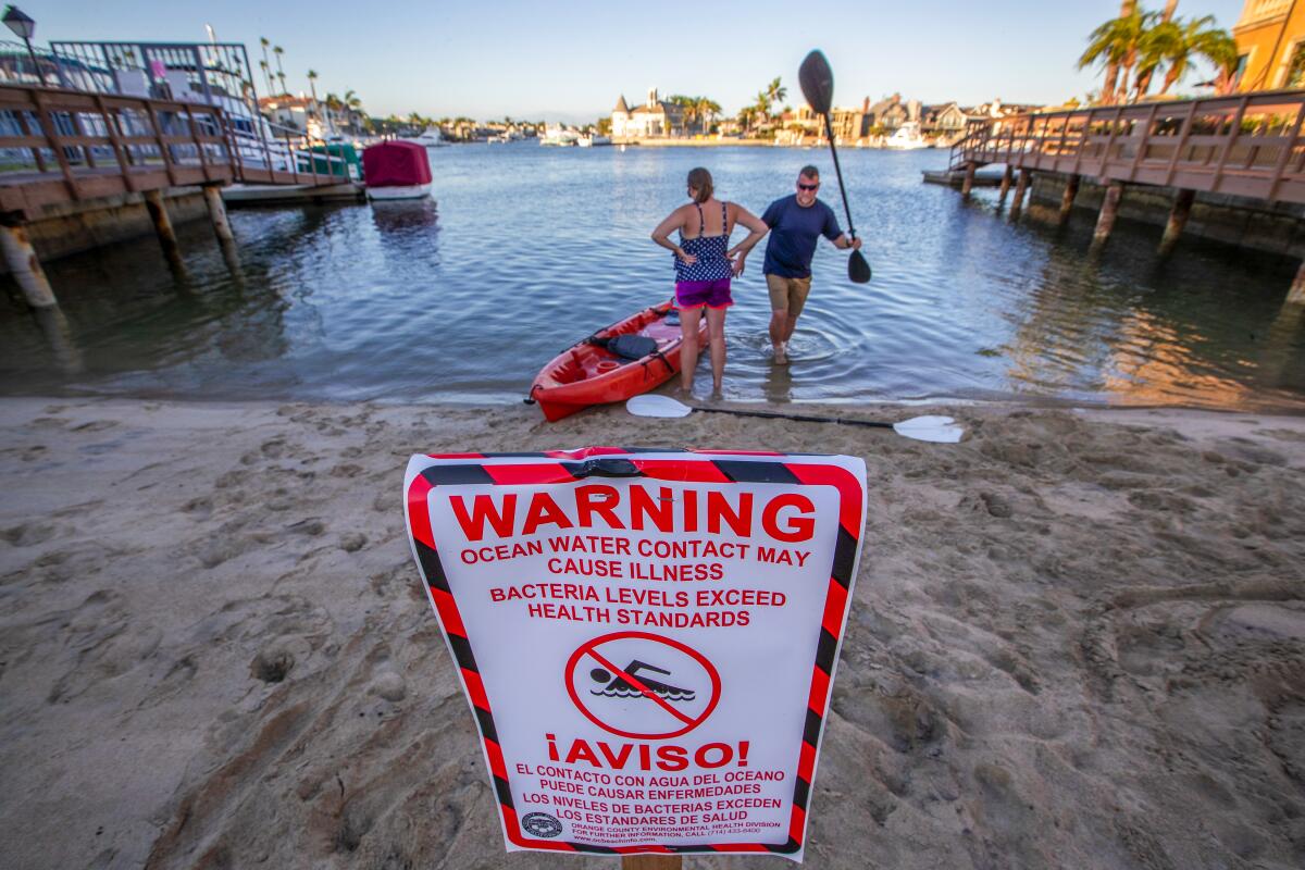 A sign in the sand warns against excess bacteria in the ocean; behind it, two people in ankle-high water with a kayak.