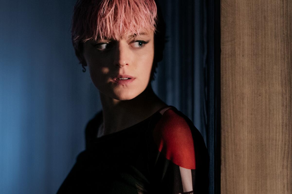 A young woman with short pink hair peers fearfully around a corner in a remote, high-tech hotel.