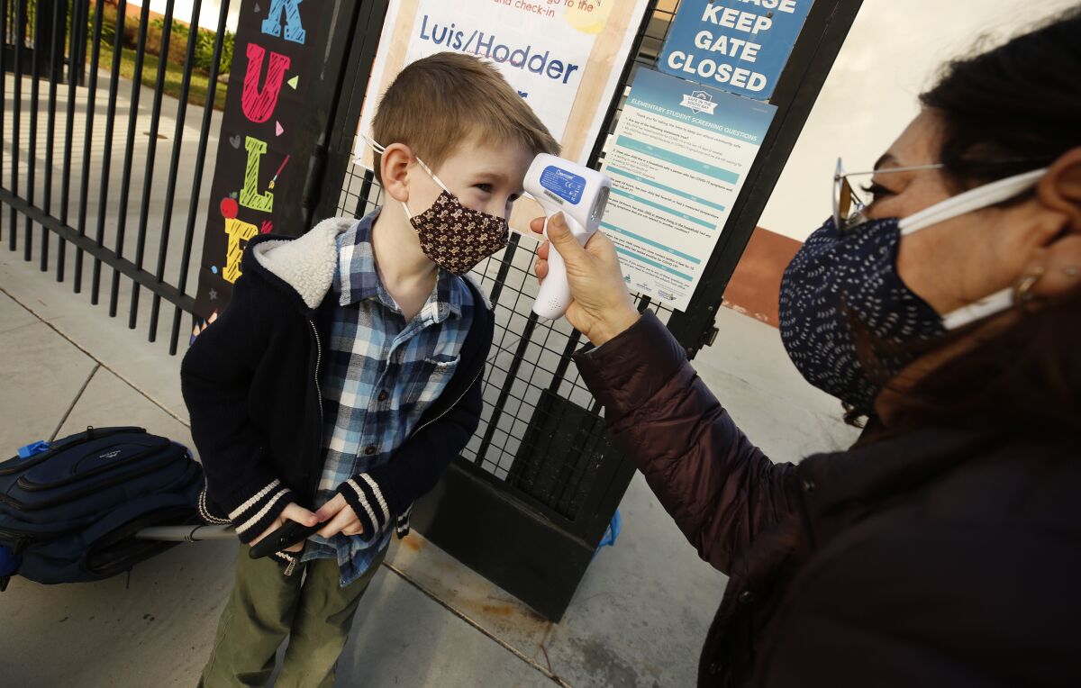 A boy rolling a backpack and wearing a mask stands as a woman holds a thermometer to his forehead.