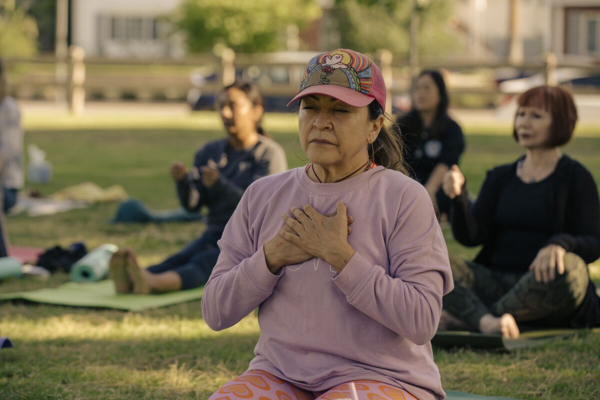 A woman sits on her knees with her hands on her heart in a park yoga class.
