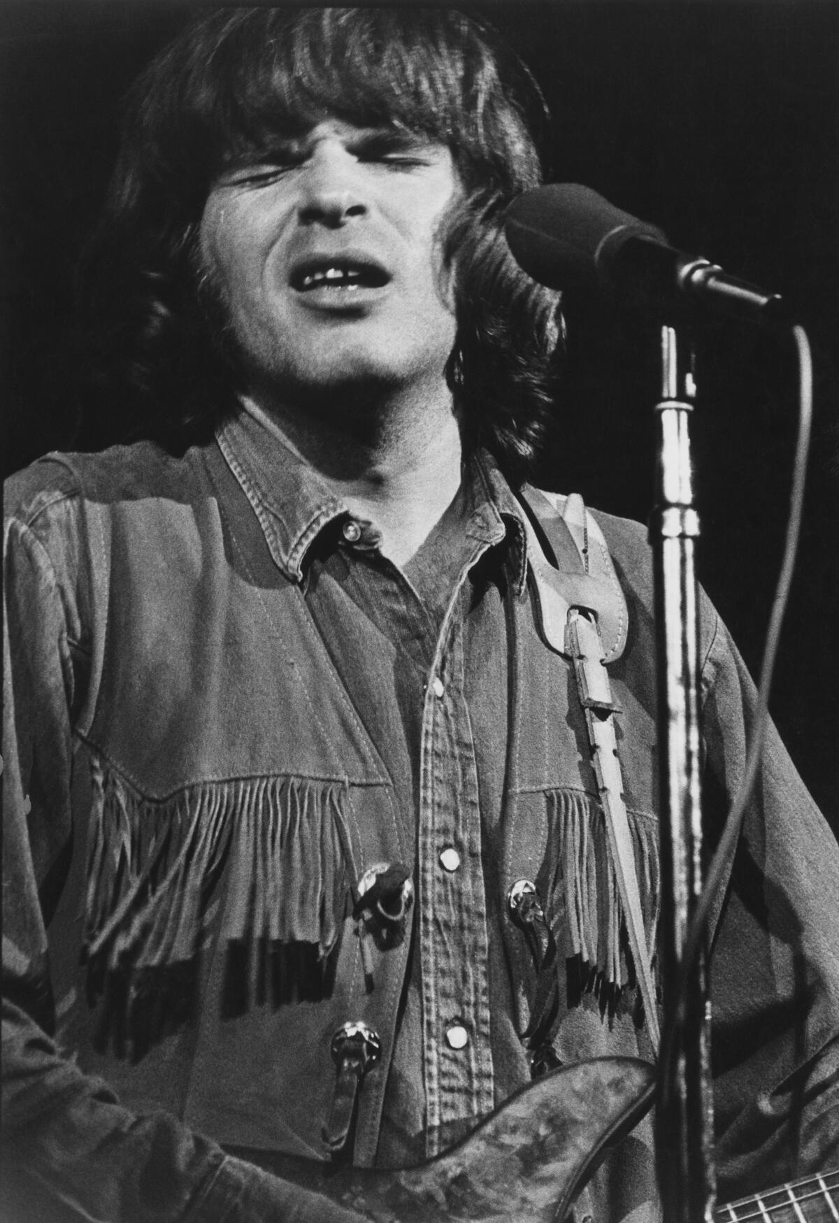 A closeup of young John Fogerty playing guitar and singing into a microphone.