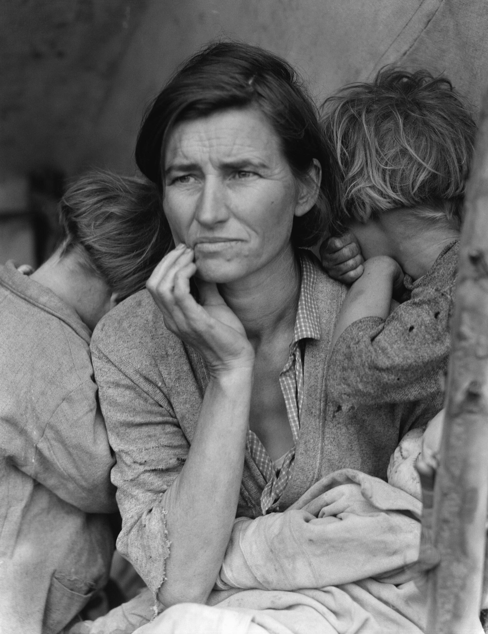 A black and white closeup of a woman with a worried expression and children leaning on her.