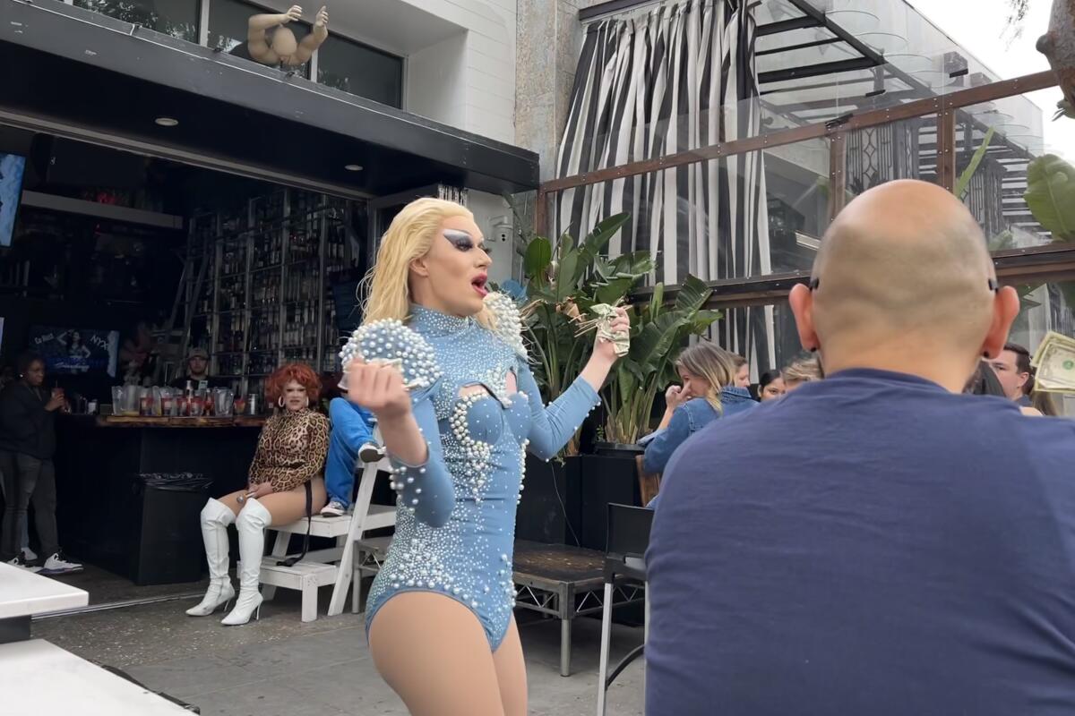 A drag queen in a blue leotard and blond wig performs on an outdoor patio