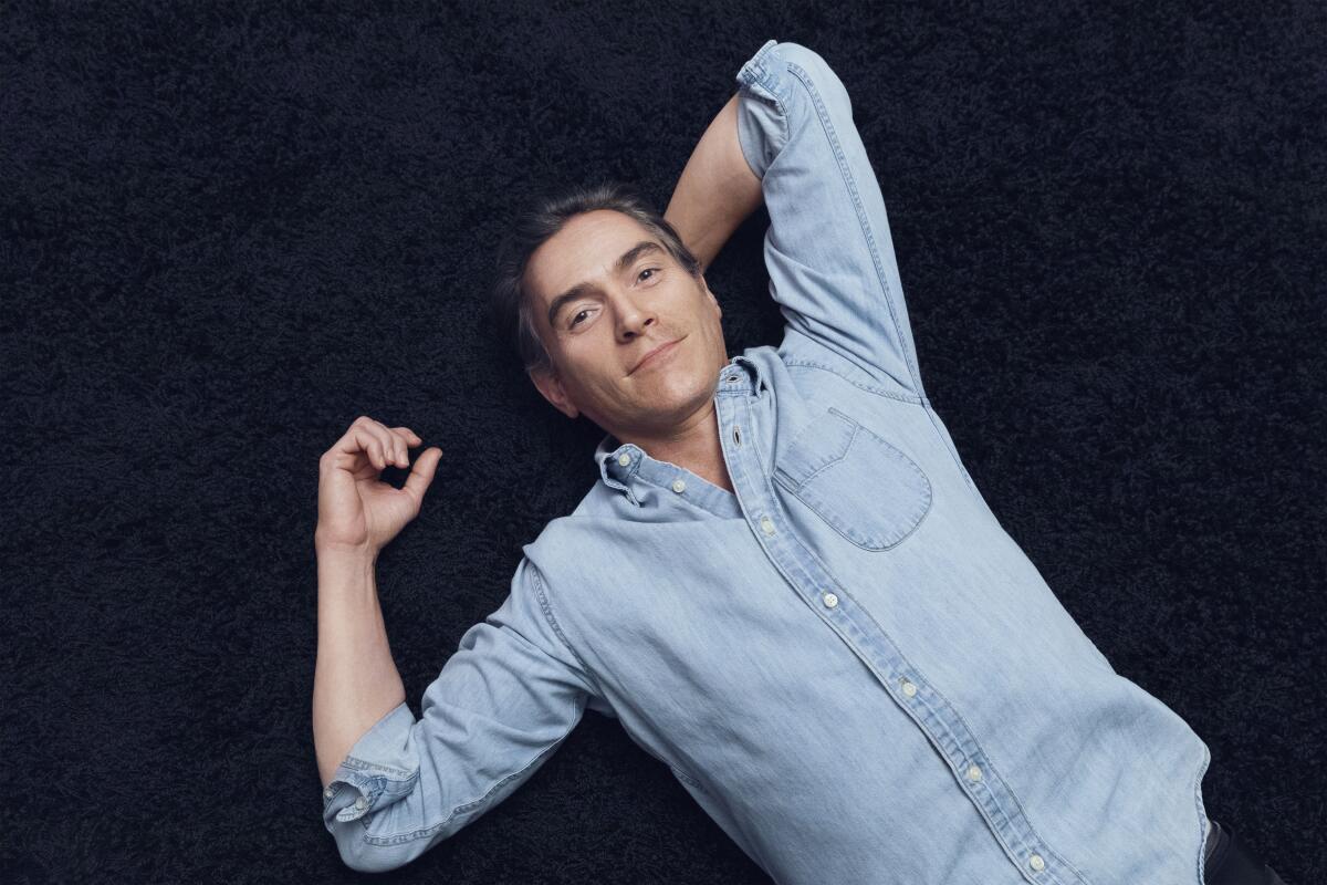 A man in a denim shirt lies on the floor, smiling up at the camera, one arm behind his head.