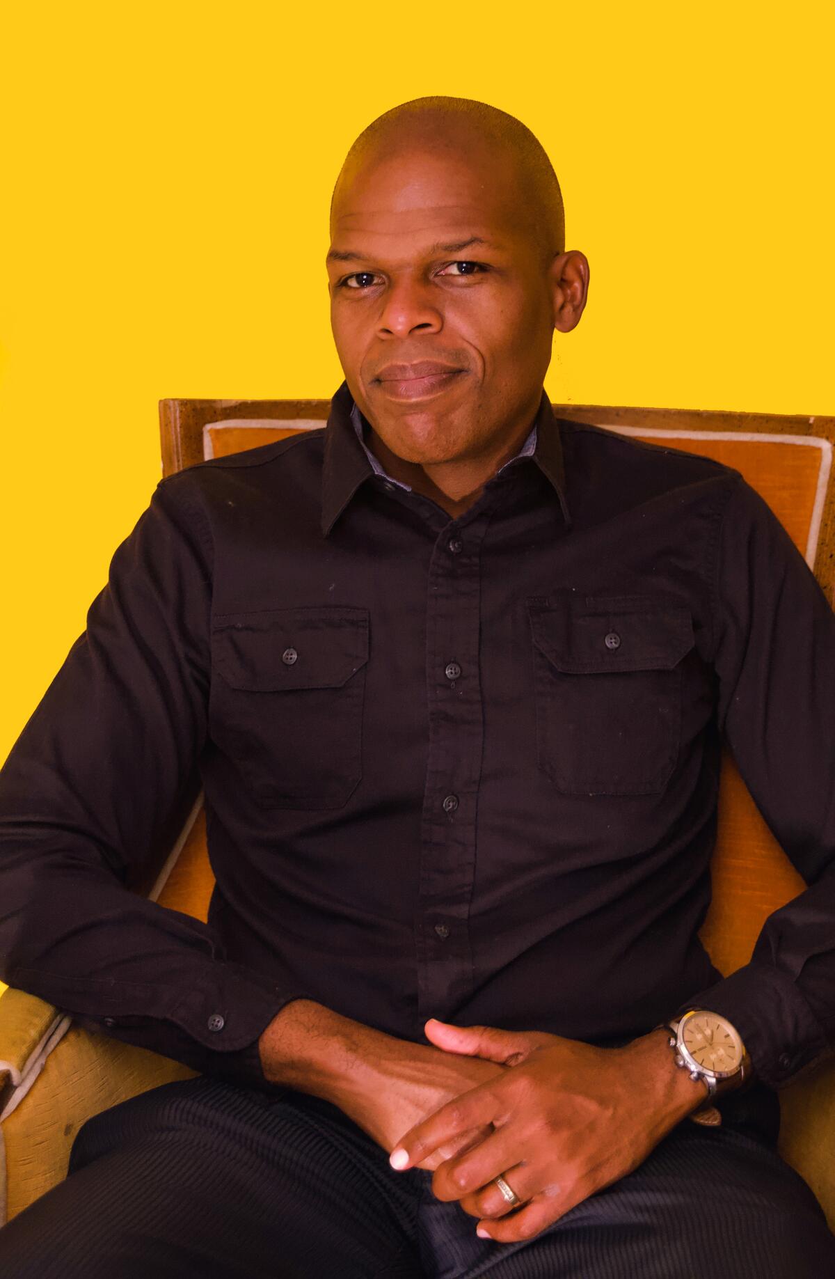 A Black man wearing all black sits in a brown chair in front of a yellow background