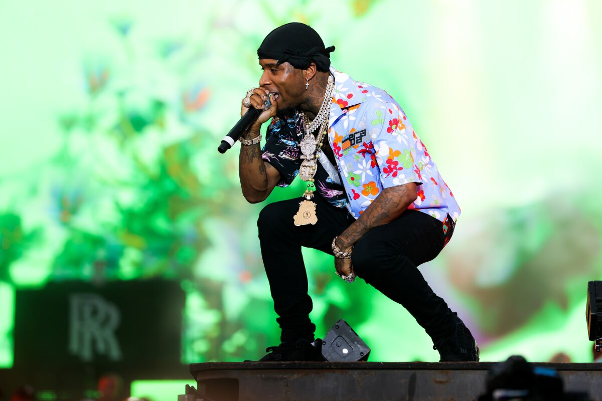 Man wearing golden chains around his neck and a blue floral shirt squats while speaking into a microphone onstage 