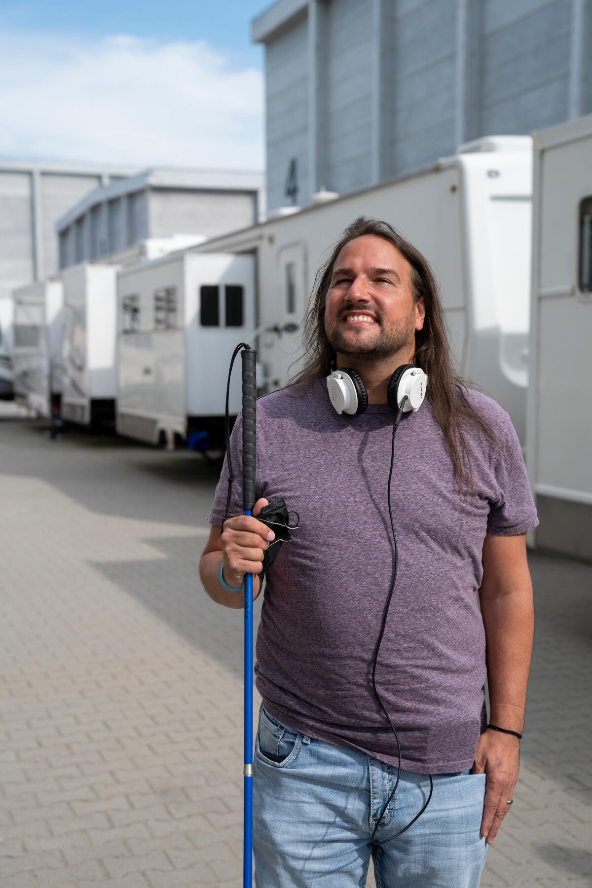 Joe Strechay stands in front of some trailers in a purple shirt and jeans with headphones around his neck and a cane in hand.