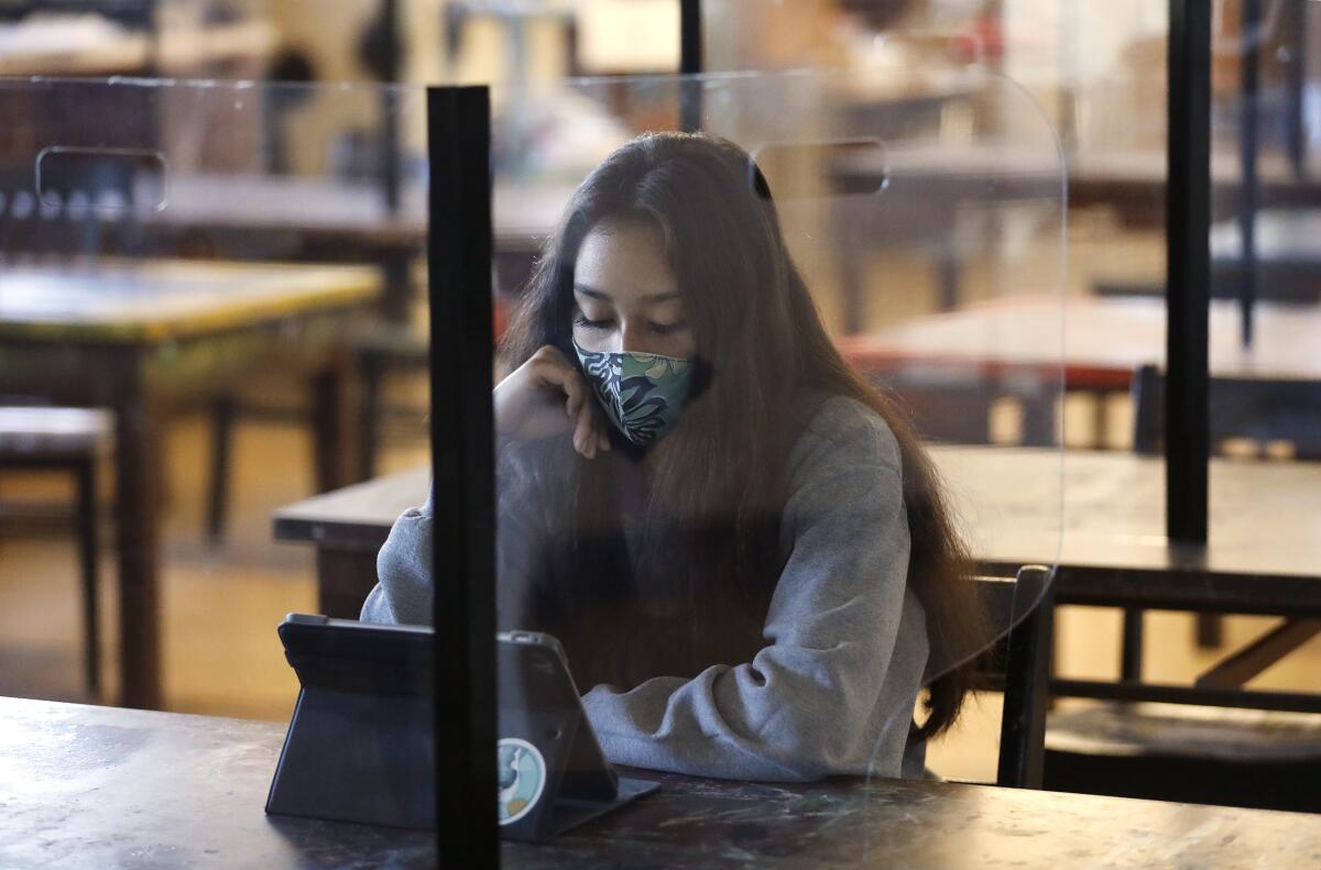 A high school student wears a mask and sits at a desk behind plexiglass.