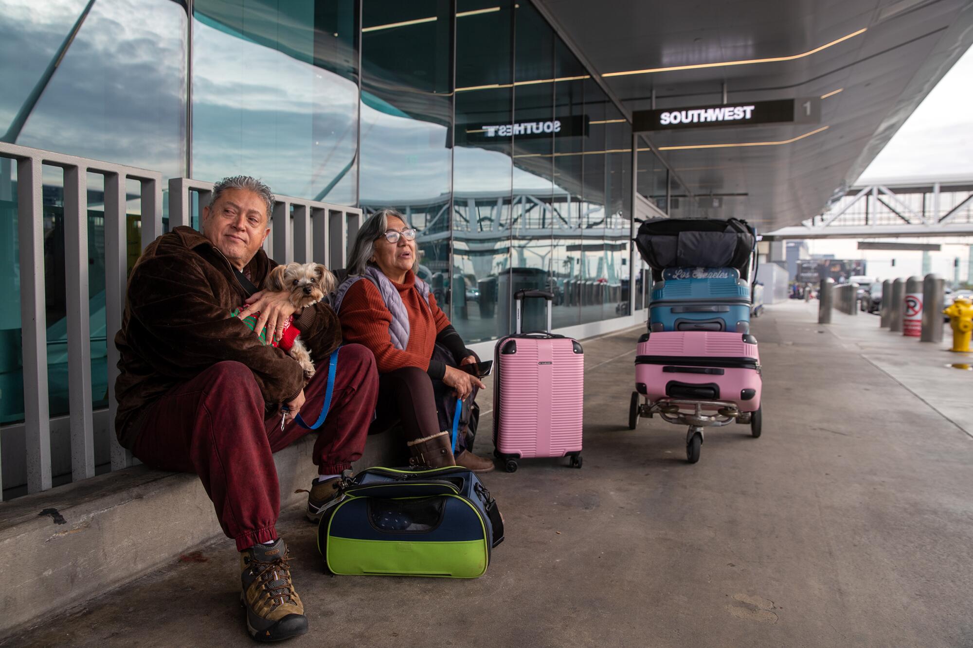 A man and a woman sit on the curb at Los Angeles International Airport with their dog and luggage.