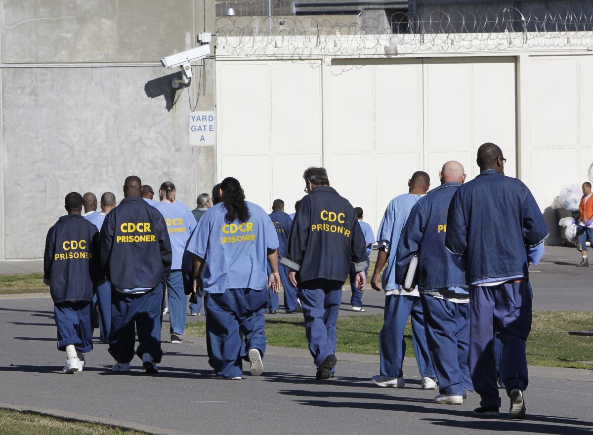 Men in blue jumpsuits with the words CDCR Prisoner on the back walk through a prison yard