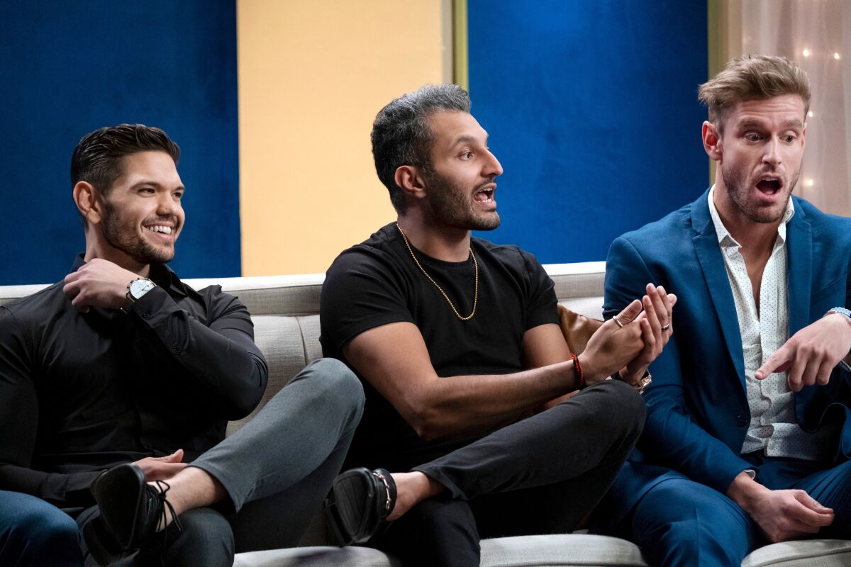 Three men on a couch looking amused, animated and shocked, respectively.