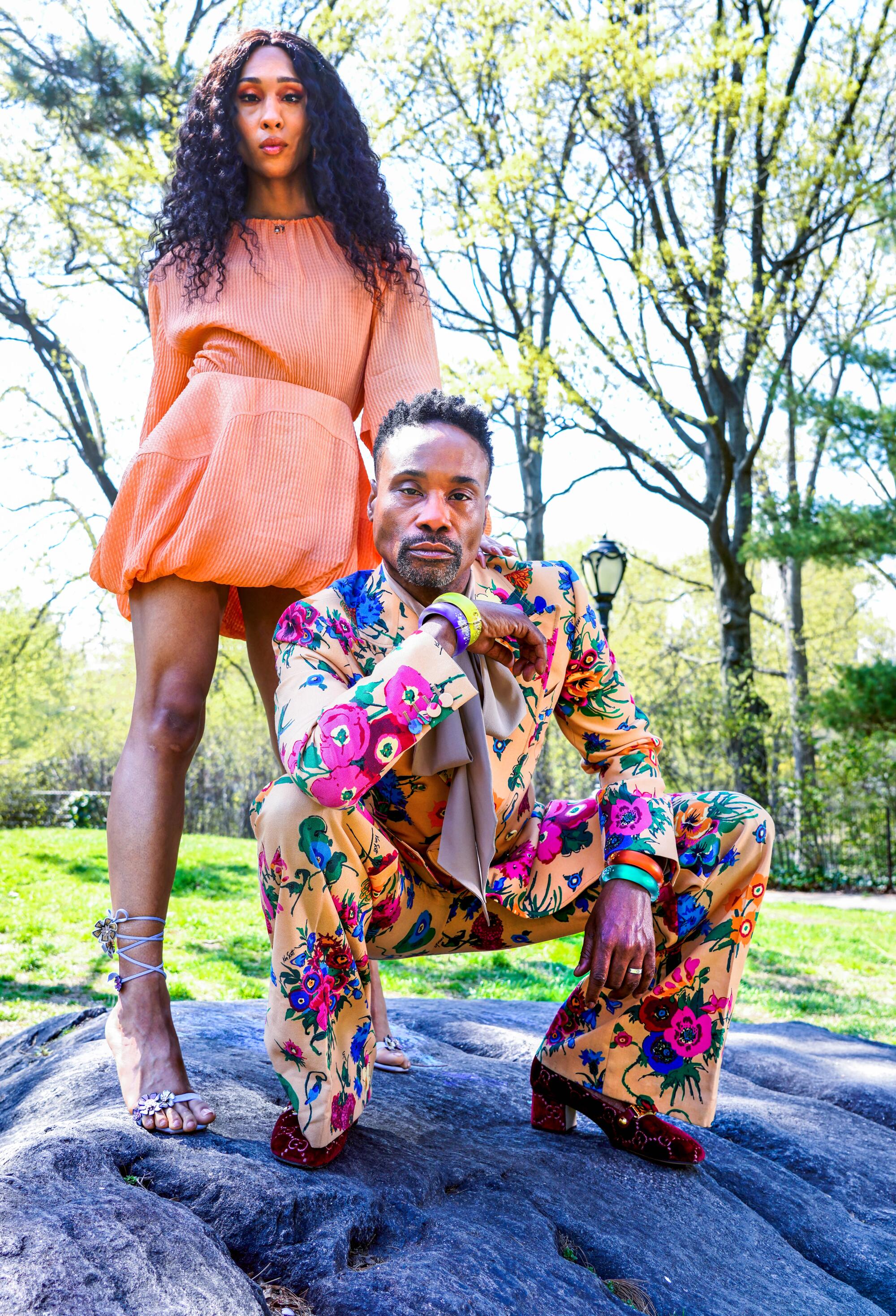 Billy Porter kneels in a flowered suit as Mj Rodriguez stands behind him in a short peach dress and sandals.