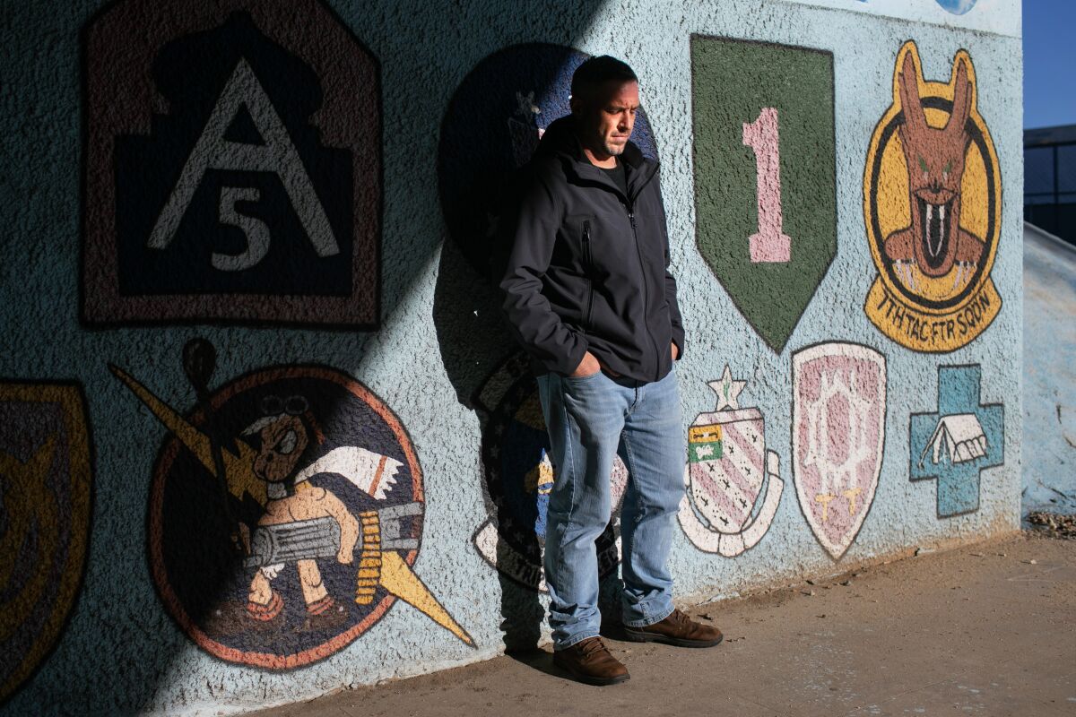 An adult man stands close to a mural displaying military units emblems. 