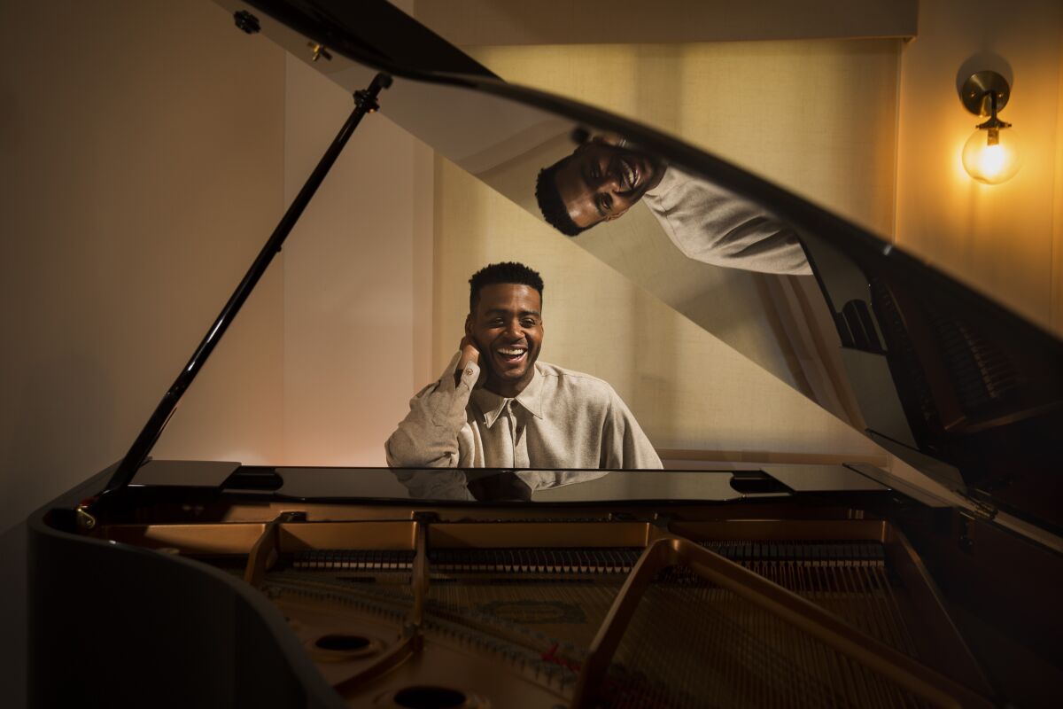 Kris Bowers, wearing a sand-colored shirt, is seen sitting at a black baby grand piano