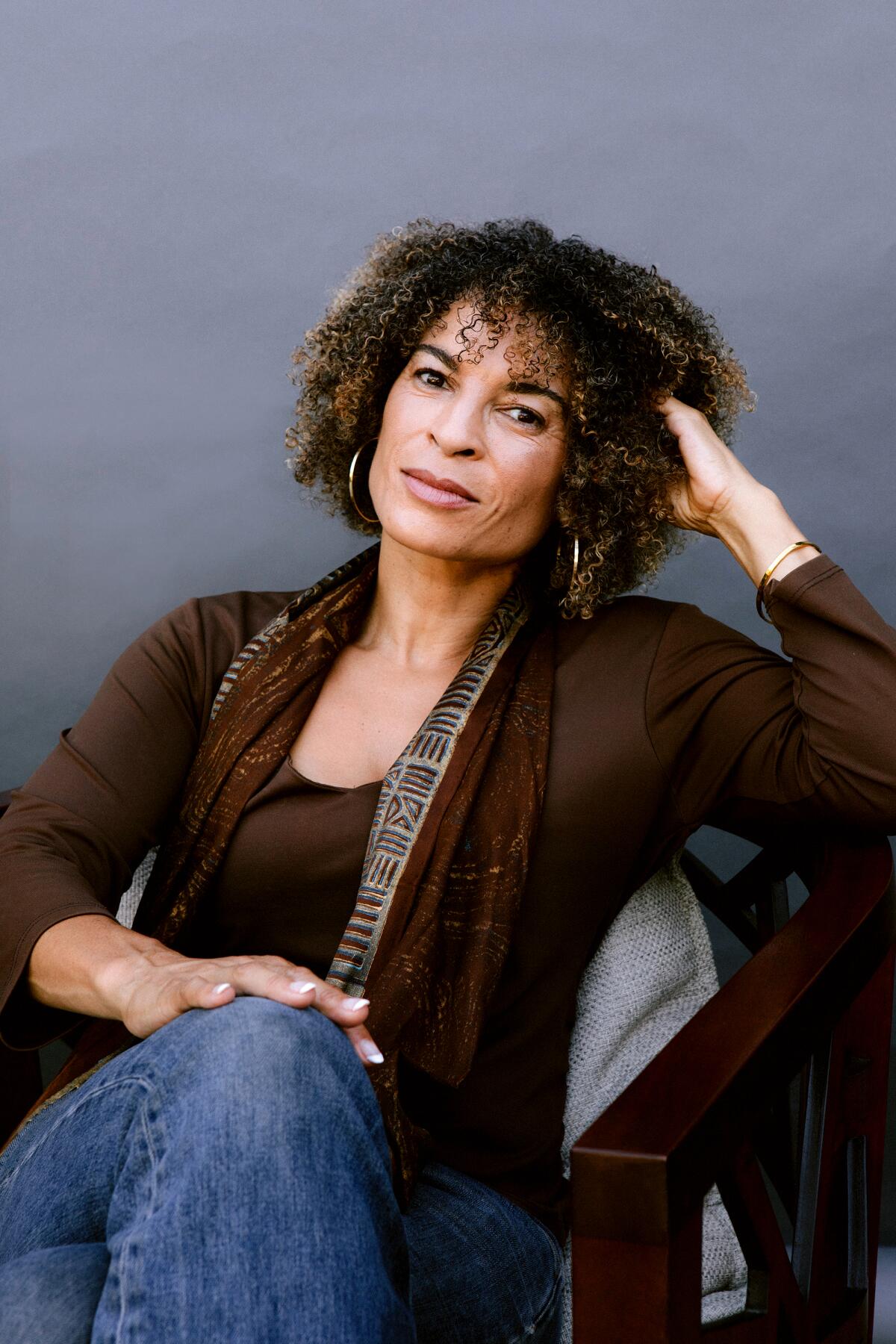 Kyra Davis Lurie, in a brown blouse and scarf, sits with one hand on her knee and the other in her hair.