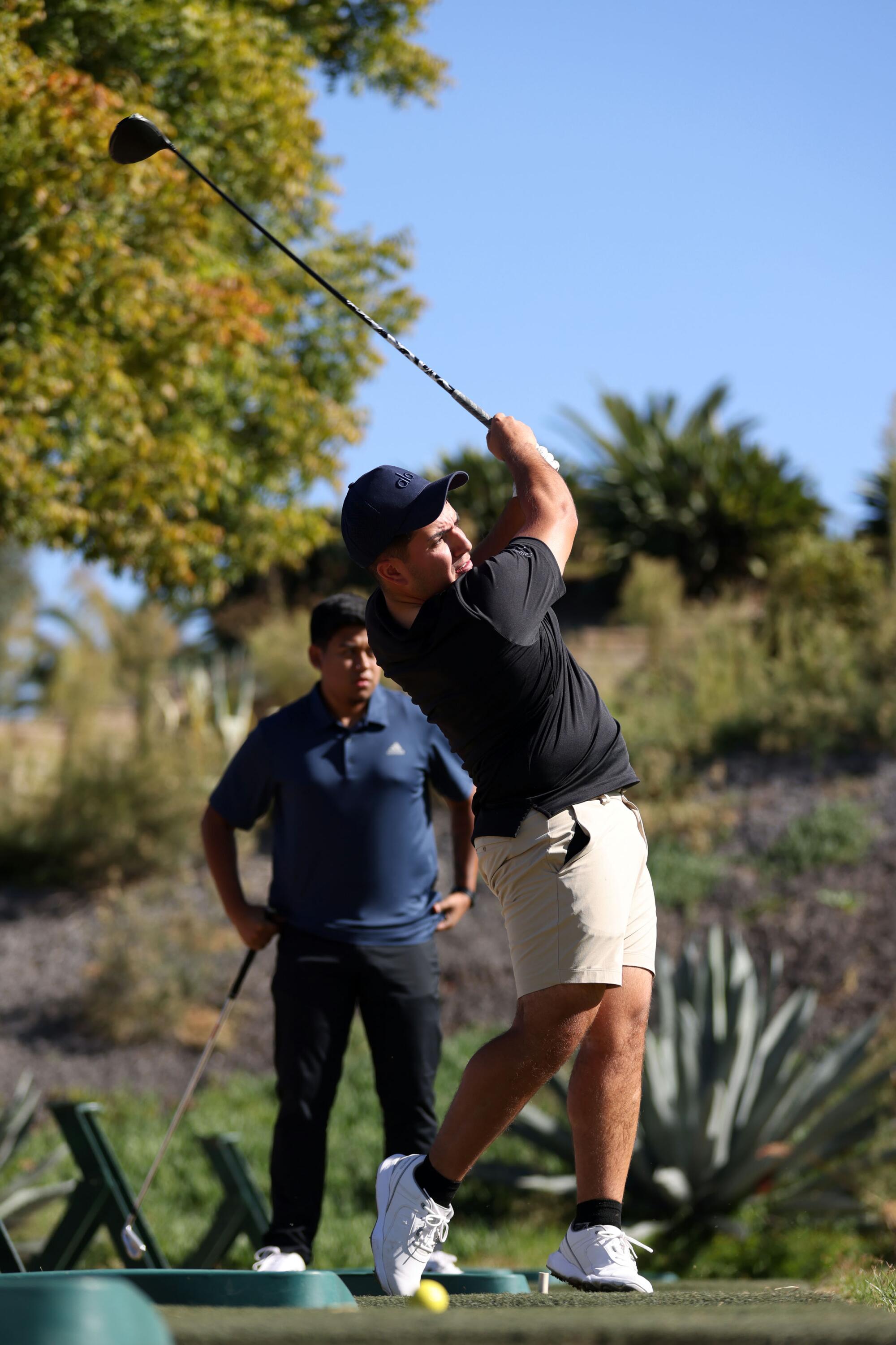 Pedro Tovar of Eslabón Armado tees off at a golf course while wearing a black shirt and white shorts.