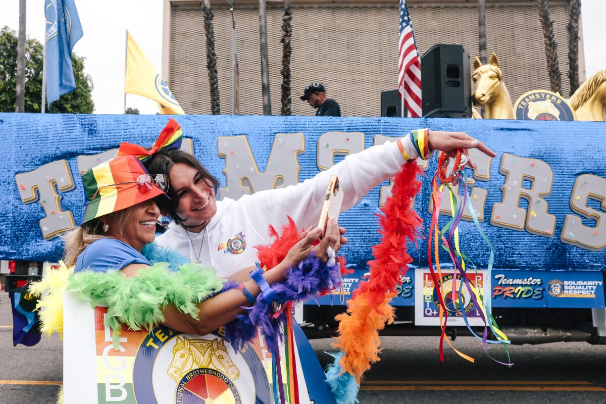 Two women dressed in rainbow outfits and accessories taking a selfie in front of a parade float on a street
