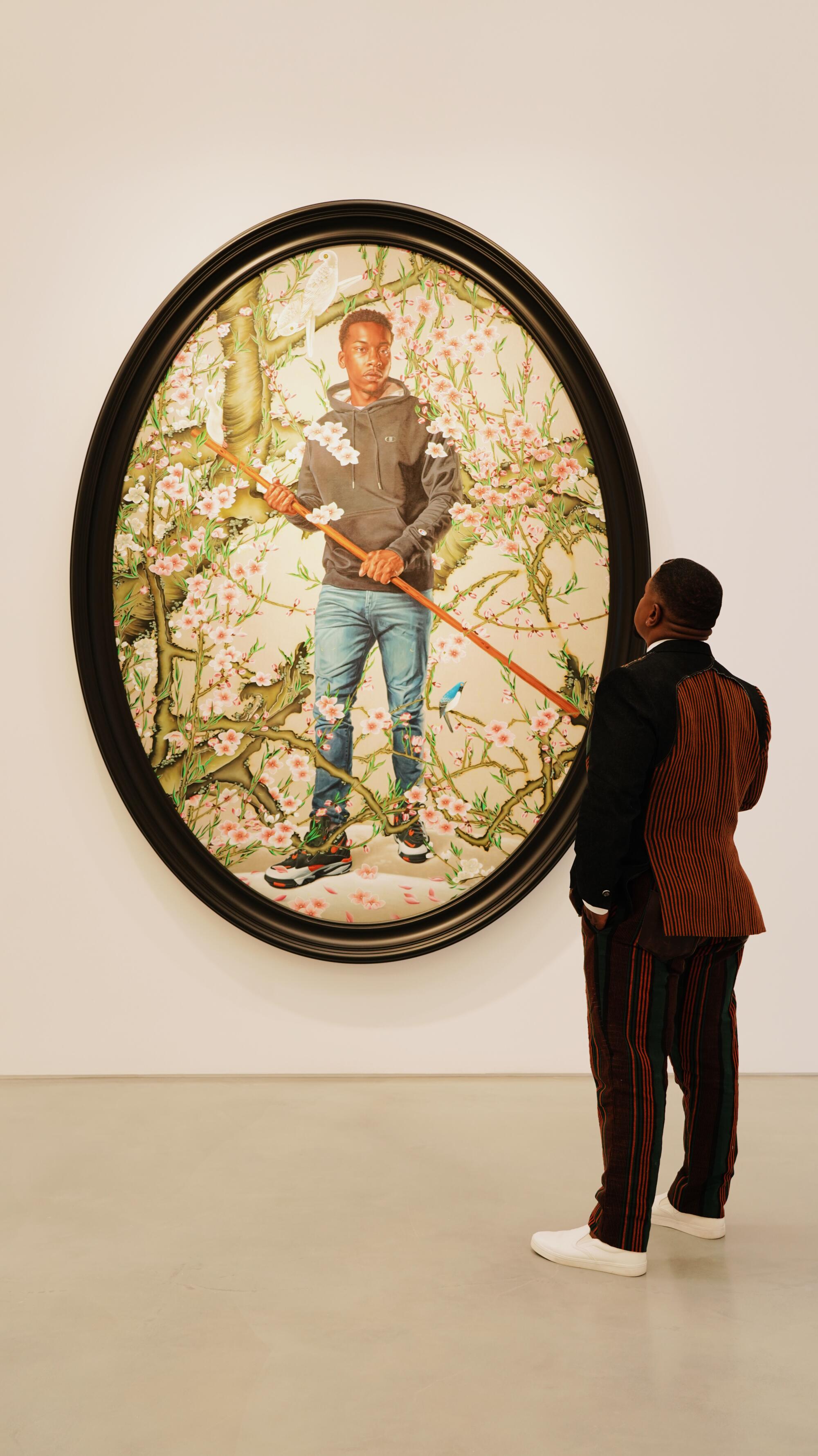 A man in a dark suit looks up at a painting of a young man in nature, in a black oval frame.