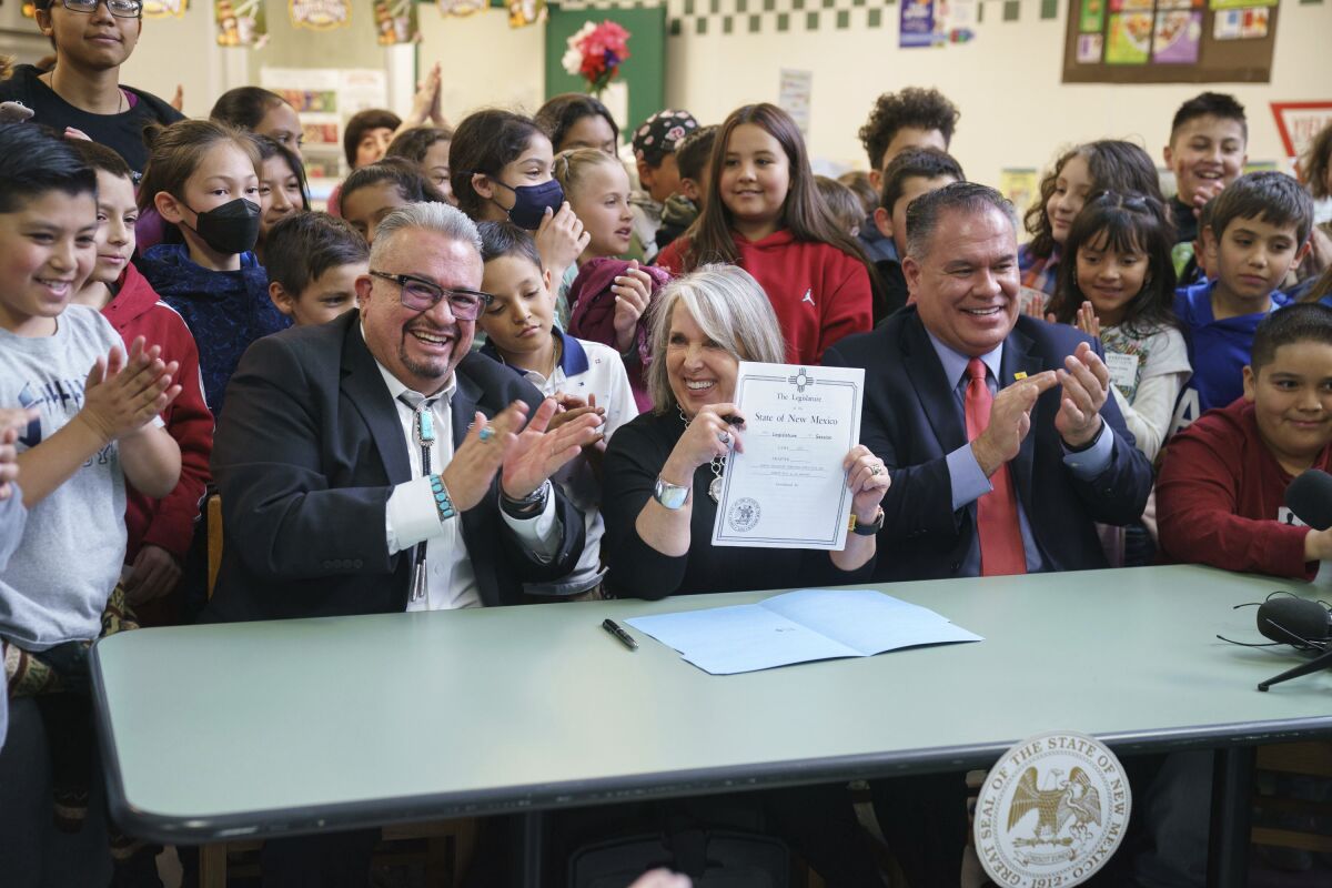 In this image provided by Gov. Michelle Lujan Grisham's office, the Democratic governor, center, holds a bill she signed during a celebration with sponsors Sens. Michael Padilla, right, and Leo Jaramillo, left, at Pinon Elementary School in Santa Fe, N.M., Monday, March 27, 2023. The legislation provides universal free school meals for New Mexico students. (David Lienemann/Office of the Governor via AP)