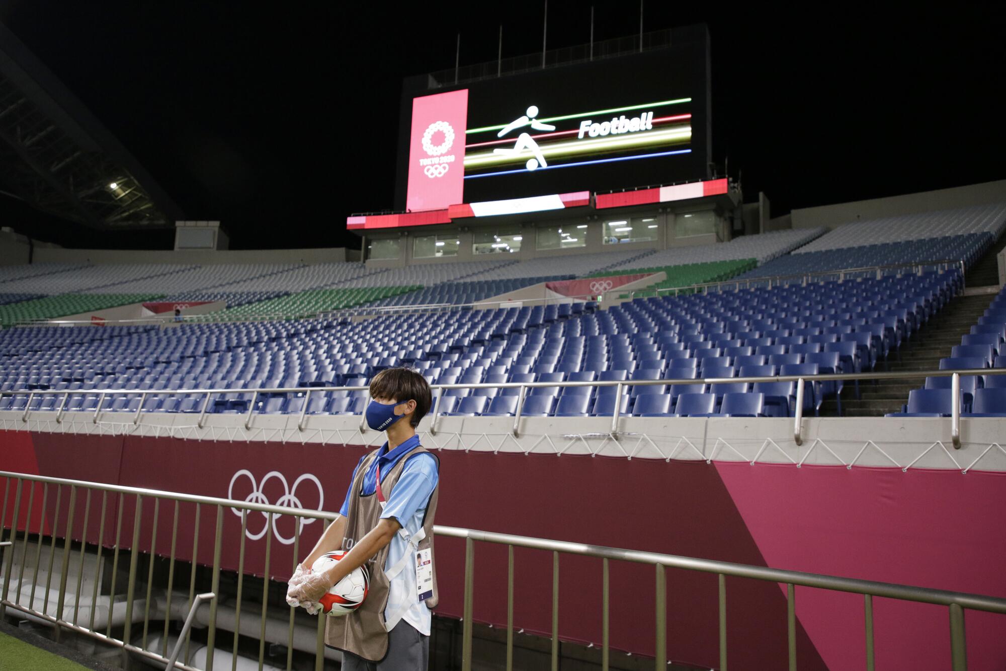 A boy holds a soccer ball. On the wall of the empty stadium behind him are the Olympic rings.