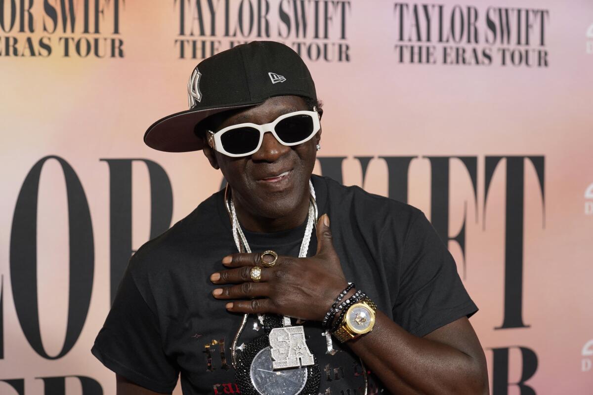 Flavor Flav, in black T-shirt and sideways black baseball cap, smiles and places a hand over his heart