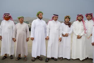Men in white robes and dark sandals, some wearing red headdresses, stand next to one another