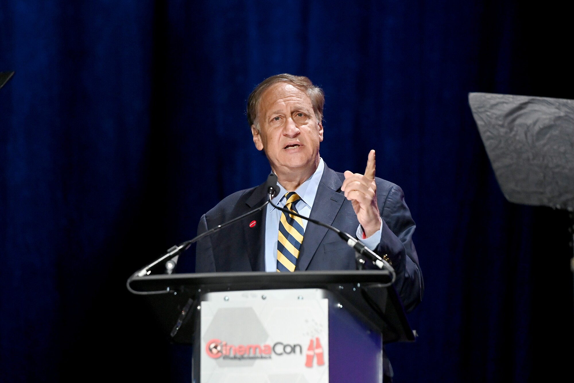 A man in a suit and tie holds up a finger as he speaks into a microphone at a podium onstage.