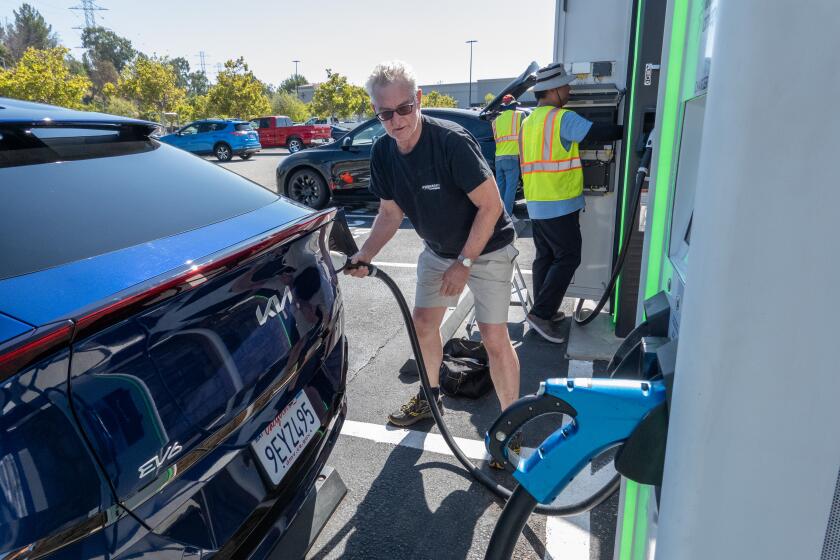 A man in a black T shirt and sunglasses attaches an electric charging pump to his blue Kia EV6 vehicle.
