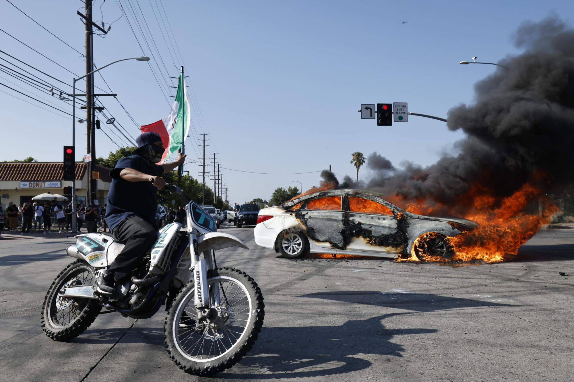 Anti-ICE protesters celebrate in front of a burning car in the middle of an intersection while a motorcyclist watches