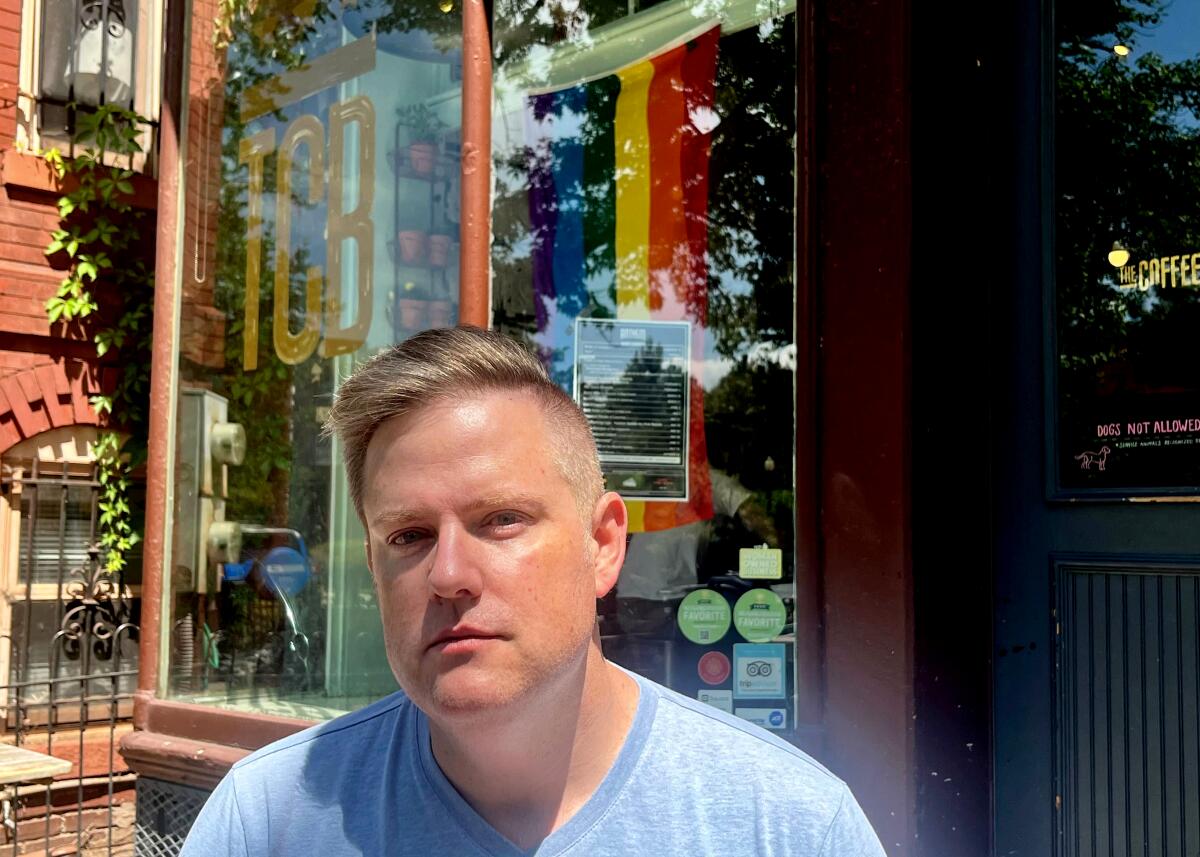 A head-and-shoulders frame of a man with short blond hair sitting in front of a coffee shop with rainbow flag in the window