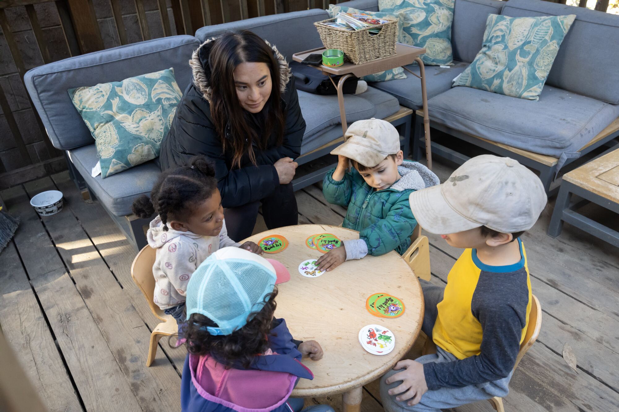 A woman in a coat sits at a small table with four children, watching as they play with animal cards.