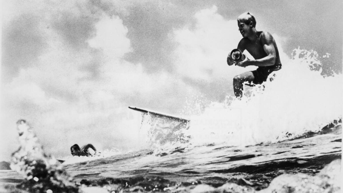 American surfer and director Bruce Brown surfs a wave and holds a camera while filming footage for his surfing documentary, "The Endless Summer."