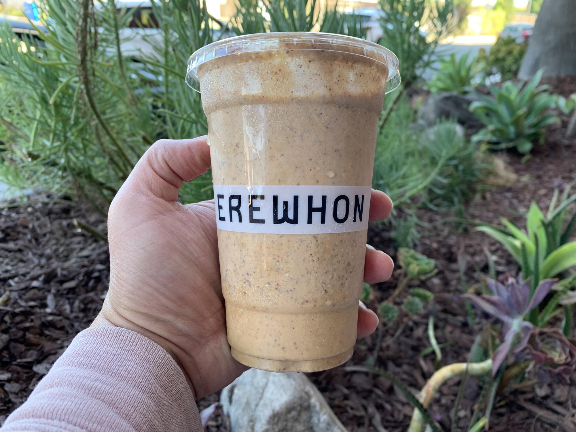 A hand holds a orangey brown smoothie in a plastic cup that says Erewhon.