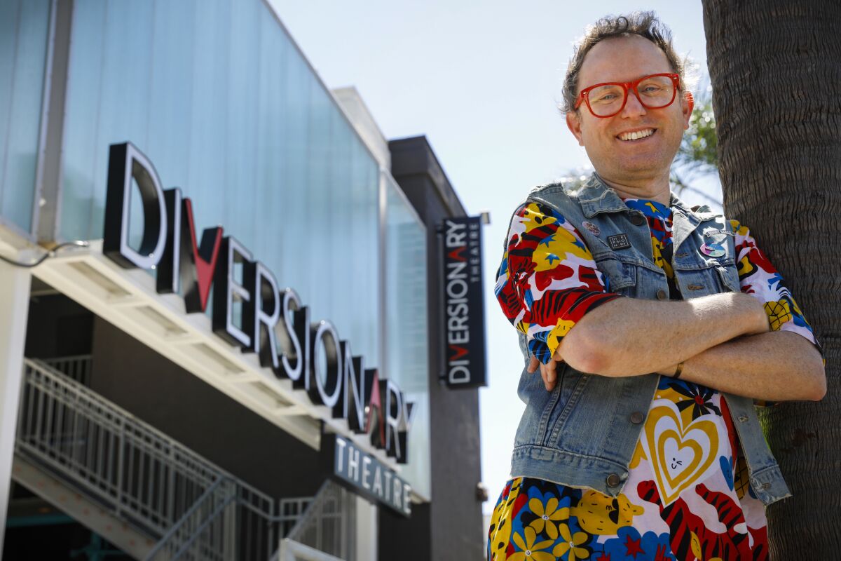 A man wearing red glasses and a denim vest over a colorful shirt poses outside Diversionary Theatre.