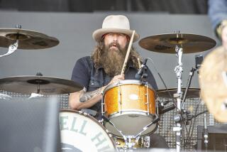 A man in a cream, wide-brimmed hat with aa bear and tattoos on his forearm sitting at and playing a drumkit on a stage