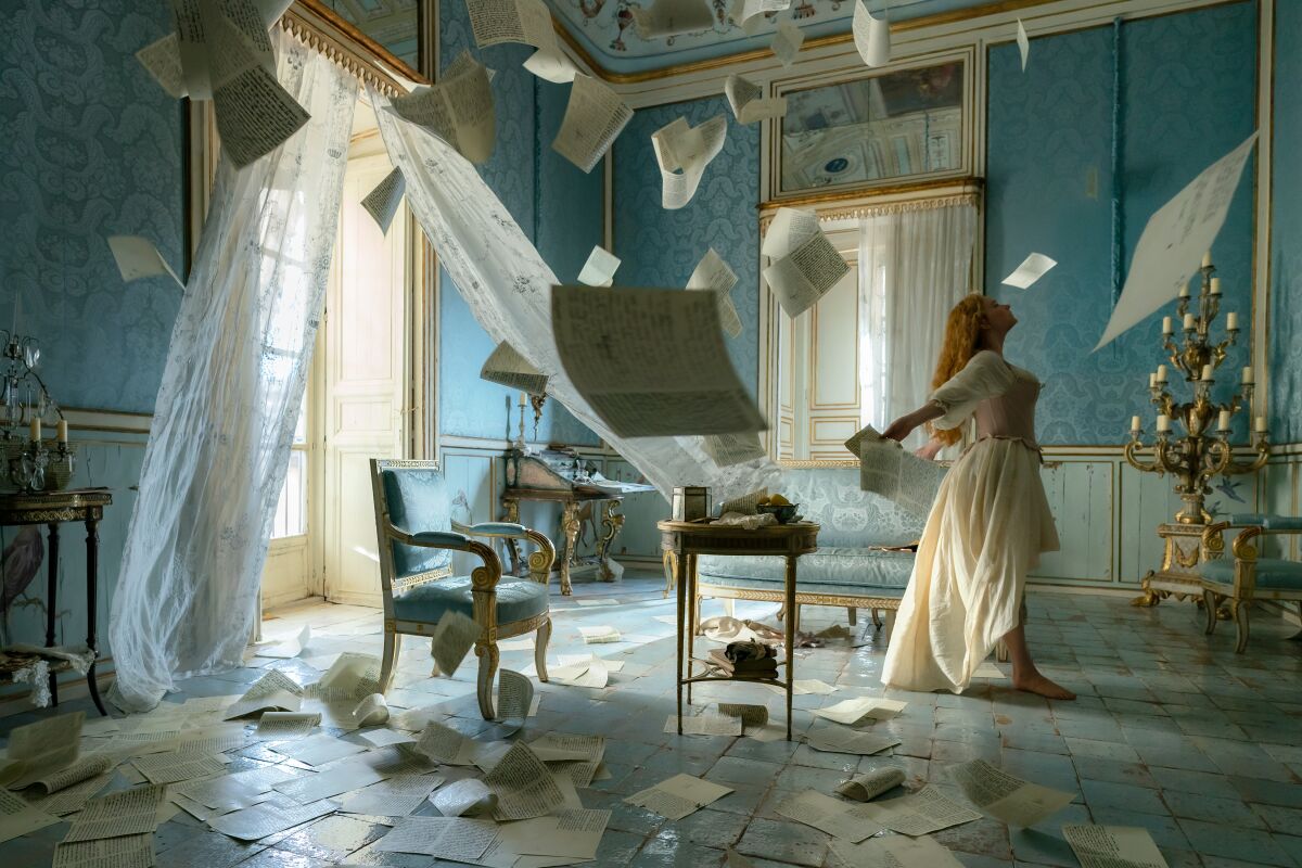 A woman stands, arms thrust back, holding a letter as the wind blows curtains and pieces of paper around a room.