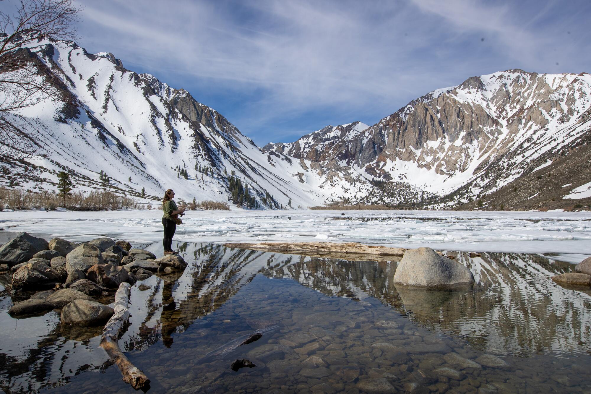 A woman casts a line in a lake surrounded by snowy mountains