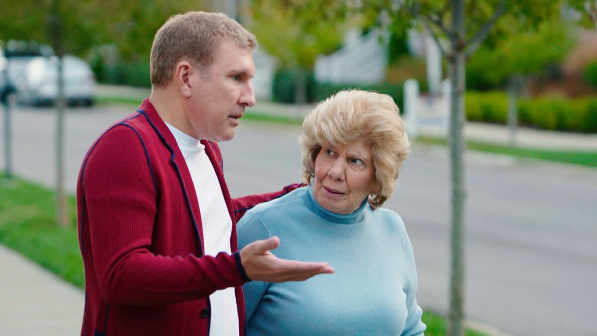 Todd Chrisley in a red cardigan holding his right hand out and standing next to his mother, Faye, in a blue turtleneck