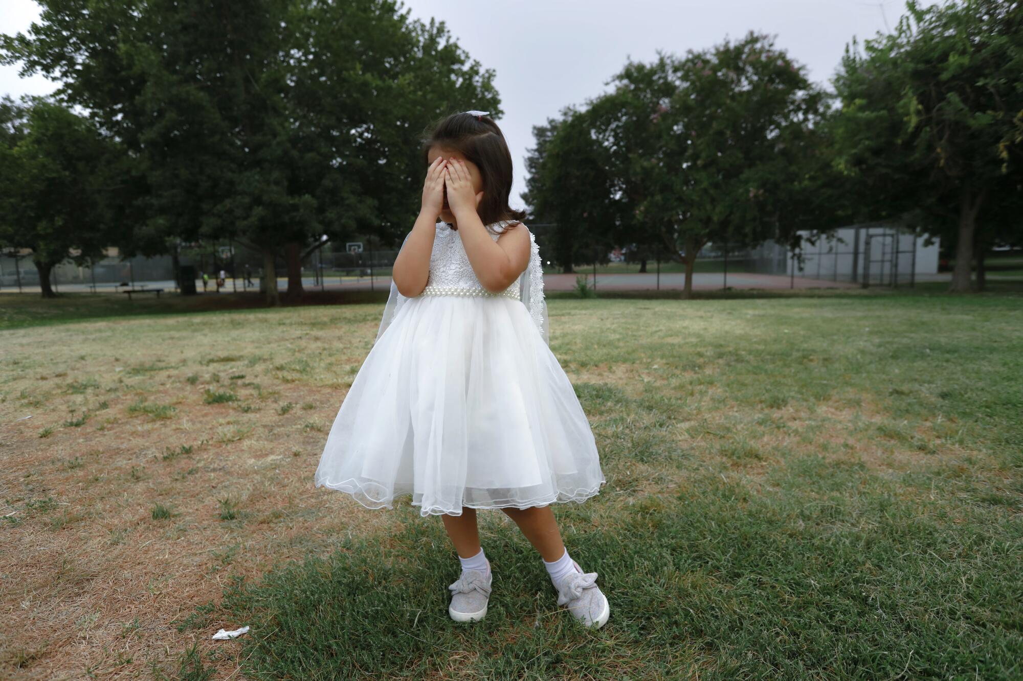 A girl in a white dress covers her eyes while playing hide-and-seek with other children at a park.