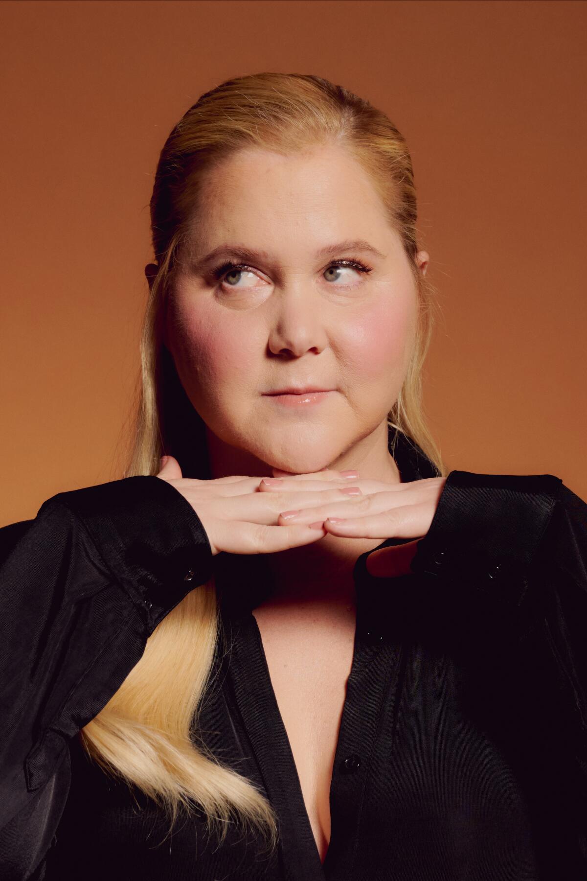 A close portrait of Amy Schumer who holds her hands below her chin and looks to the side.