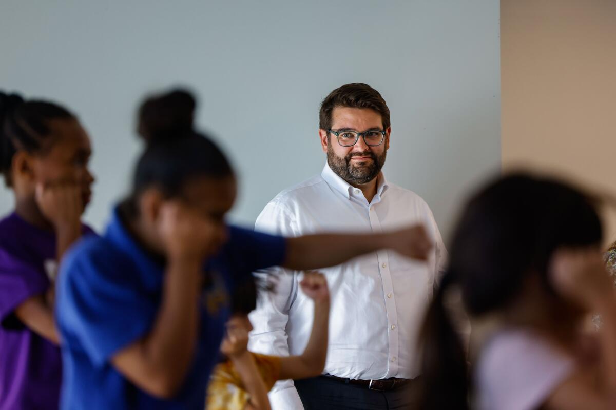A bearded man with dark hair, wearing glasses and a white dress shirt, looks at children in boxing stances
