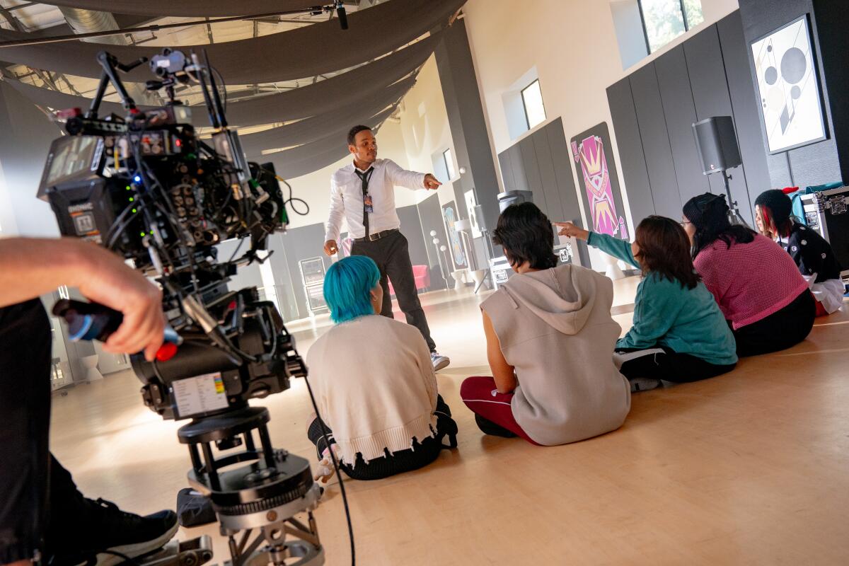 A man directs five people seated on the floor in front of him. A movie camera stands nearby.