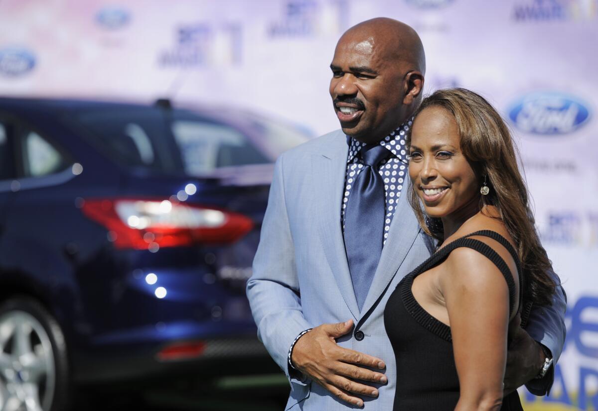 Steve Harvey in a suit and tie and Marjorie Harvey in a black dress smile and pose together in front of a car