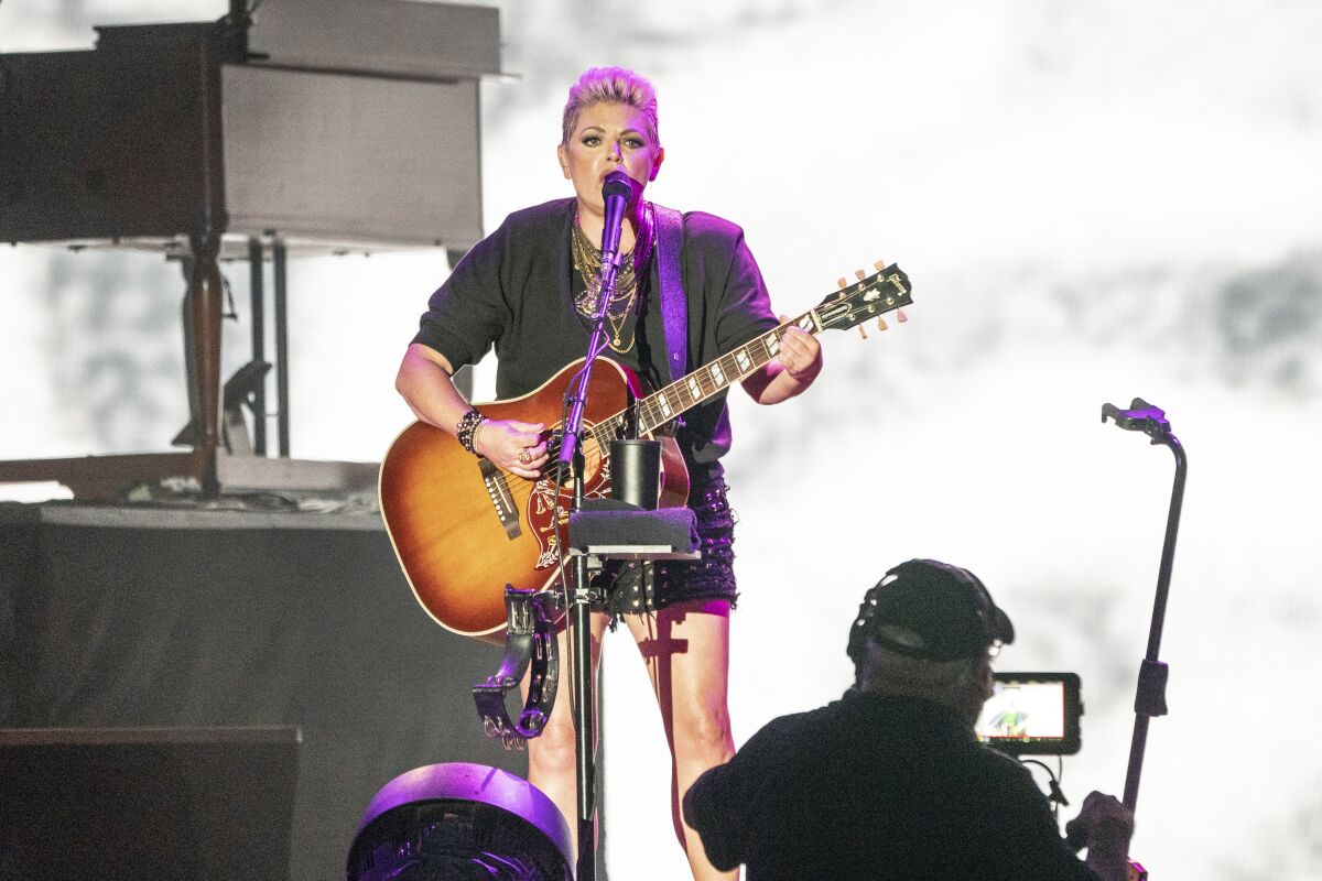 A woman with short blond hair stands onstage playing a guitar